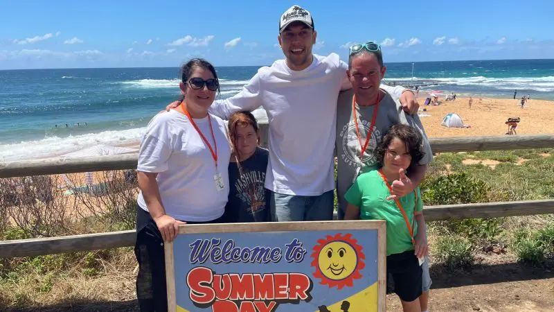 Six happy tourists, including two children, pose by the “Welcome to Summer Bay” sign on a sunny beach during a 1 Day Home and Away Tour.