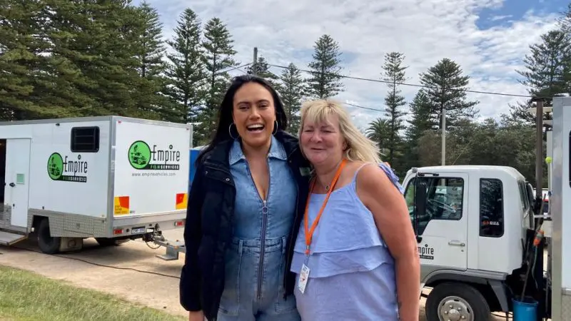 Two women smiling beside lorries with tall pine trees in the background, filming a 1 Day Home Away Tour by Celebtime Tours for fans.