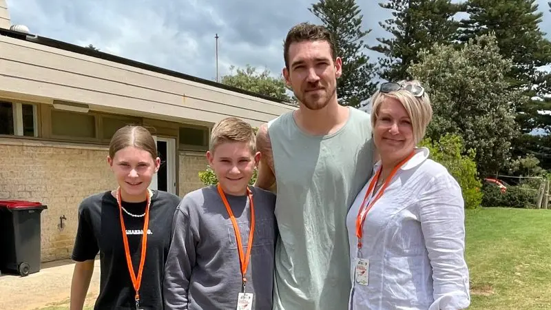 Group of four wearing orange lanyards stands outdoors on a 1 Day Home Away Tour, smiling by a building surrounded by green trees.