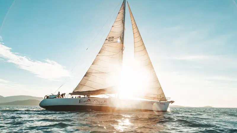 Experience the Broomstick 2 Day 2 Night Whitsunday Sailing boat gliding at sunset, golden sunlight streaming through sleek white sails.