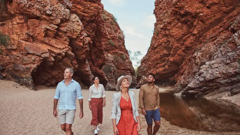 Group of four walkers exploring towering red rock cliffs on a 1 Day West MacDonnell Ranges & Standley Chasm tour in Australia.