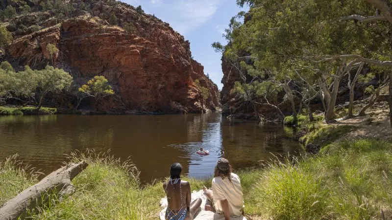 Two travellers relax beside a tranquil river, admiring vibrant red cliffs on a guided 4-Day Uluru to West MacDonnell tour, blue sky overhead.