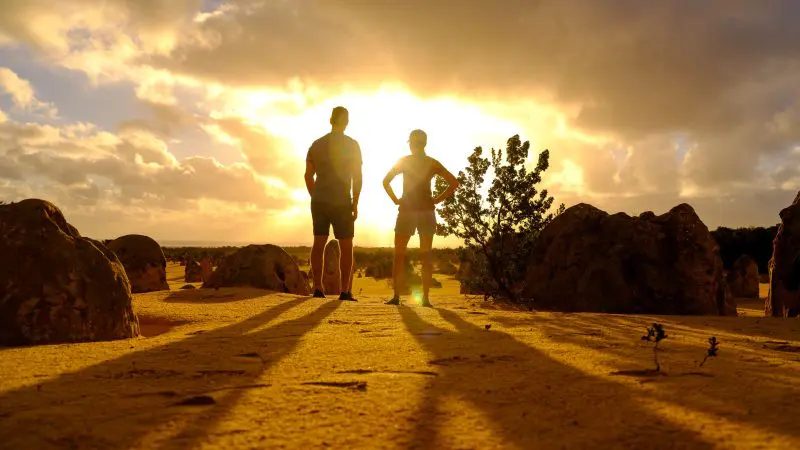 Two people on sandy terrain at sunset during a 1 Day Pinnacles Sunset Stargazing Tour Autopia, with vibrant clouds behind them.