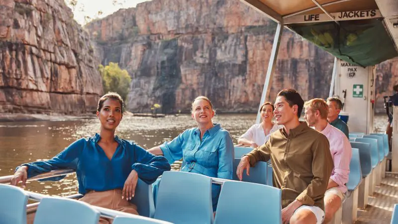 Six happy tourists on a 1 Day Katherine Gorge Cruise admire dramatic sandstone cliffs and sparkling water beneath a bright sunny sky.