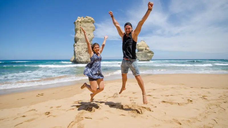 Two happy travellers jump on a pristine beach during the 3 Day Great Ocean Road & Grampians Explorer Melbourne Return tour adventure.