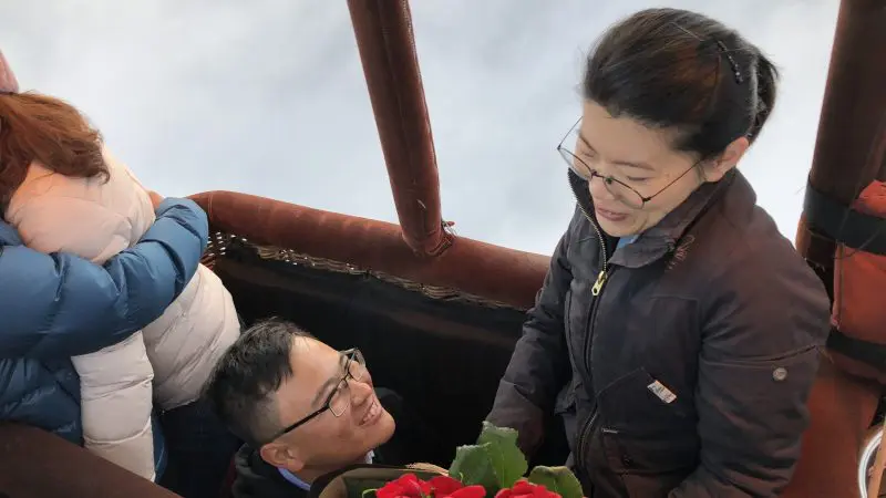Romantic hot air balloon proposal over Melbourne City, man holding red roses above clouds with skyline views in breathtaking sunrise.