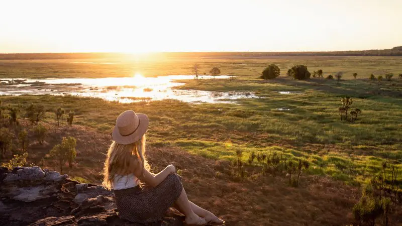 Woman wearing a hat sits on a rock, watching sunset on a 5 Day Kakadu, Katherine Gorge & Litchfield 4WD tour from Darwin.