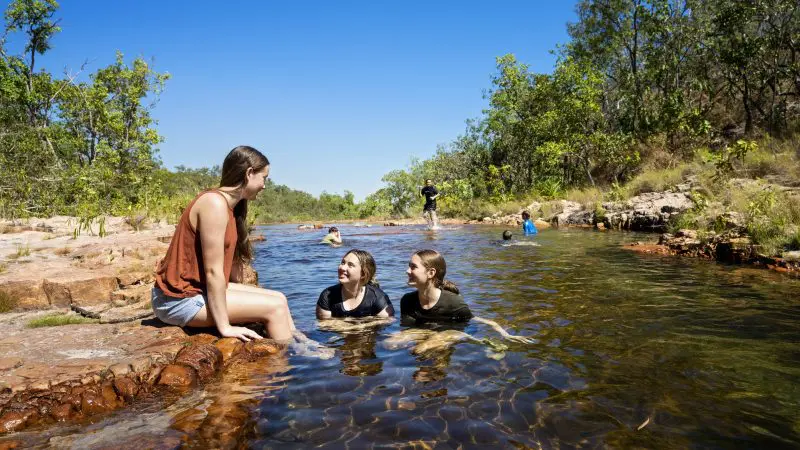 Three people unwind in a crystal-clear creek, surrounded by lush greenery at Litchfield National Park under a vibrant blue sky, 1-day tour.