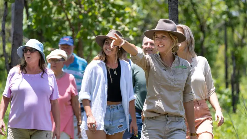 Tour group on 1 Day Litchfield National Park Crocodile Cruise, led by woman guide pointing ahead surrounded by lush green trees.