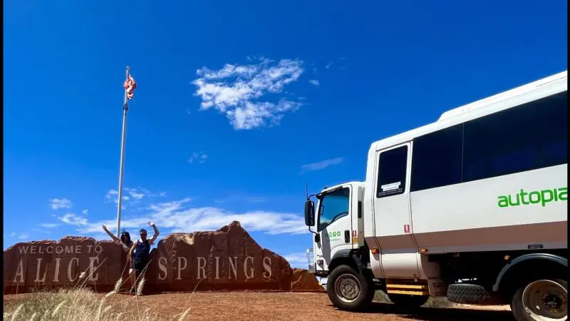 Travellers smile next to a Welcome to Alice Springs sign and Red Centre Rock Tour bus, ready for their 2-day Outback adventure.