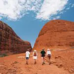 Experience four adventurers exploring iconic red rock formations on a 4-day Uluru, Kata Tjuta & Kings Canyon tour beneath clear blue skies.