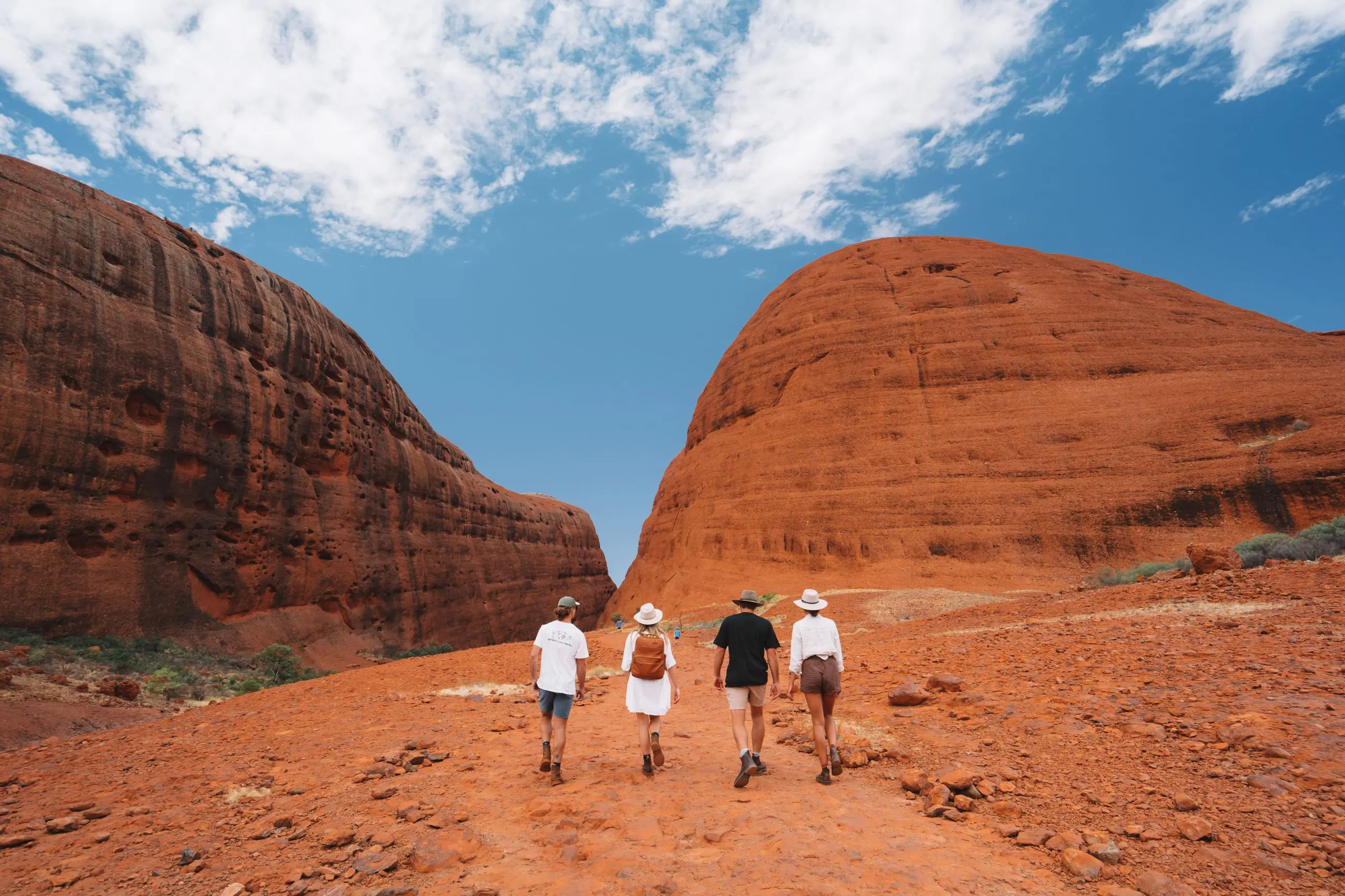 Experience four adventurers exploring iconic red rock formations on a 4-day Uluru, Kata Tjuta & Kings Canyon tour beneath clear blue skies.