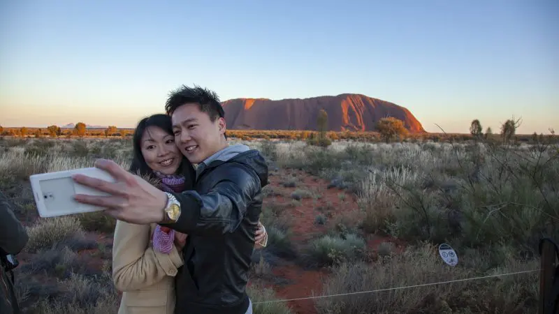 A smiling couple snaps a sunset selfie at Uluru on their 2 Day Uluru Kata Tjuta Rock Tour from Ayers Rock Yulara, iconic outback view.
