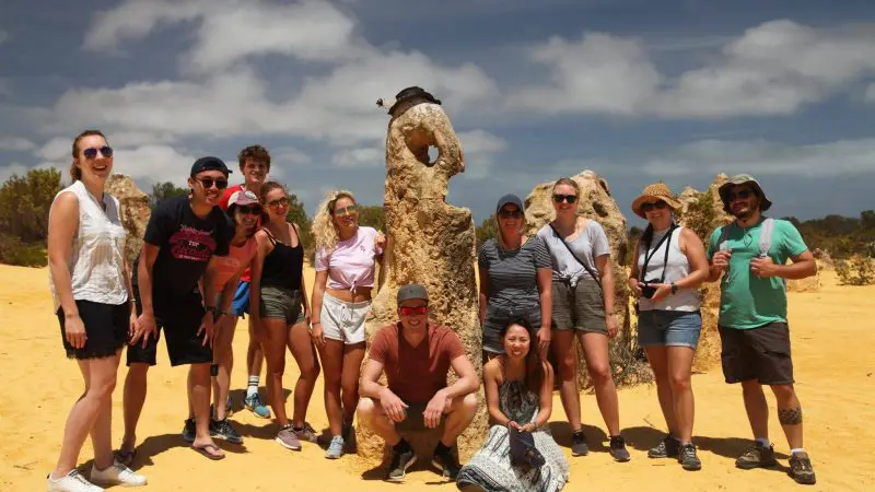 Twelve happy travellers pose by a towering rock in nature, enjoying the 6 Day Perth to Exmouth Coral Coaster adventure tour.