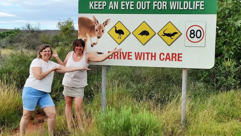 Two adventurous women smiling and pointing at a wildlife warning sign on their 6 Day Perth to Exmouth Coral Coaster tour in Australia.