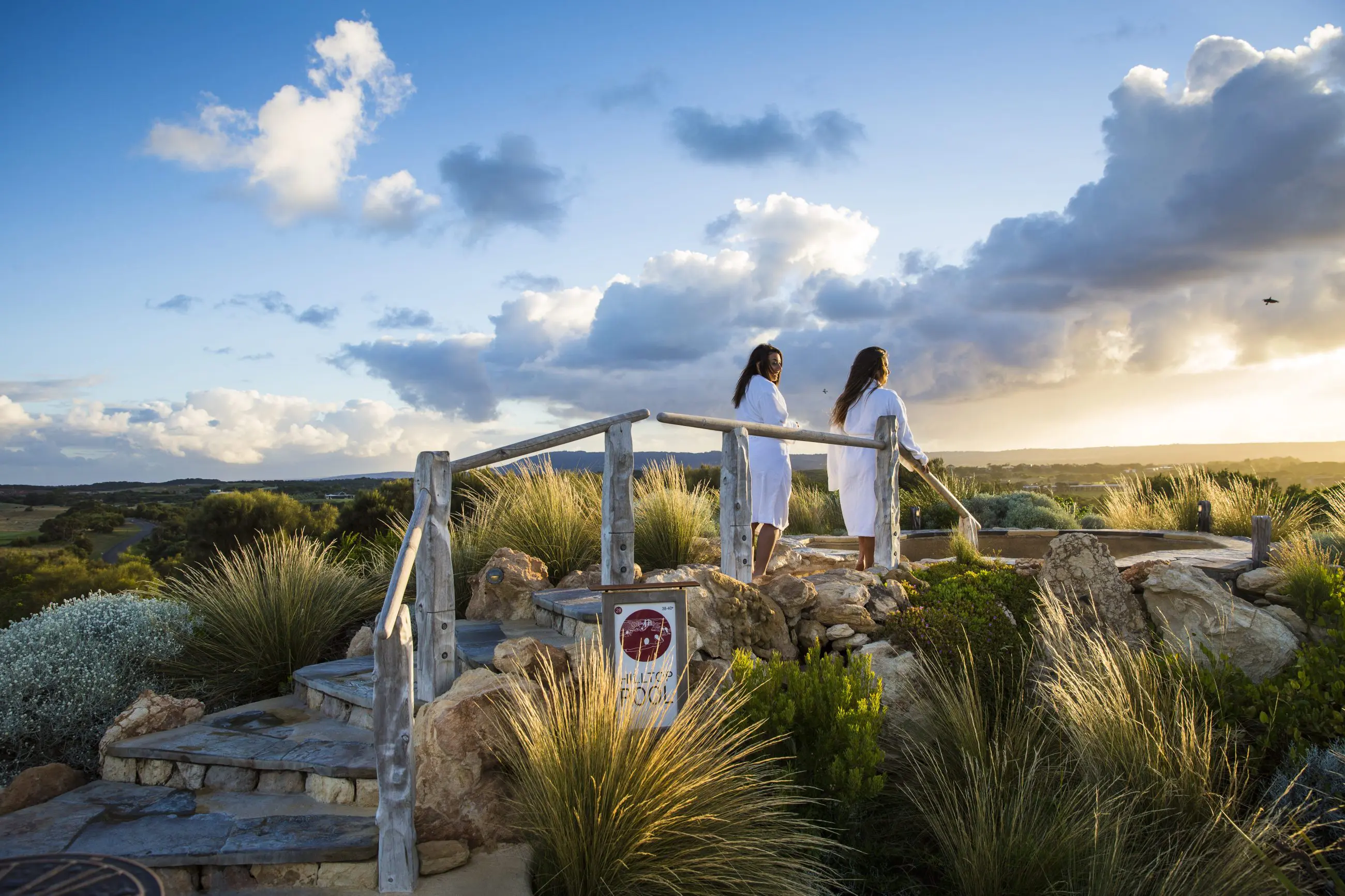 Two women in white spa robes relax at sunset after a 1 Day Peninsula Hot Springs Spa Entry Shuttle Bus experience, serene backdrop.
