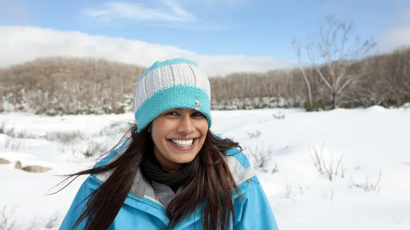 Happy woman in blue jacket enjoying a 1 Day Lake Mountain Snow Waterfalls Tour from Melbourne, with scenic snowy trees behind her.