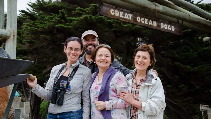 Group of four happy adults stand beneath the iconic Great Ocean Road sign on a 1 Day Great Ocean Road Rainforest Tour in Australia.