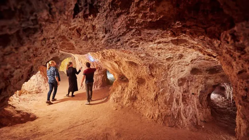 Three adventurers explore a vast orange-brown cave during a 7 Day Uluru to Adelaide Tour with Untamed Escapes, Australia.