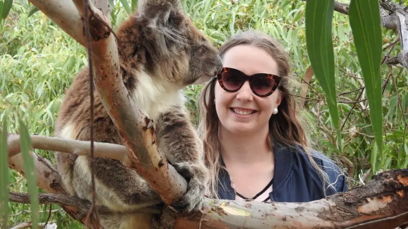 Woman in sunglasses grins beside a koala in a tree on her 10-Day Perth to Adelaide Adventure Tour—unforgettable wildlife encounter.