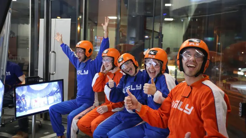 Group of five in helmets and flight suits smiling together at an iFLY Family and Friends indoor skydiving adventure experience session.