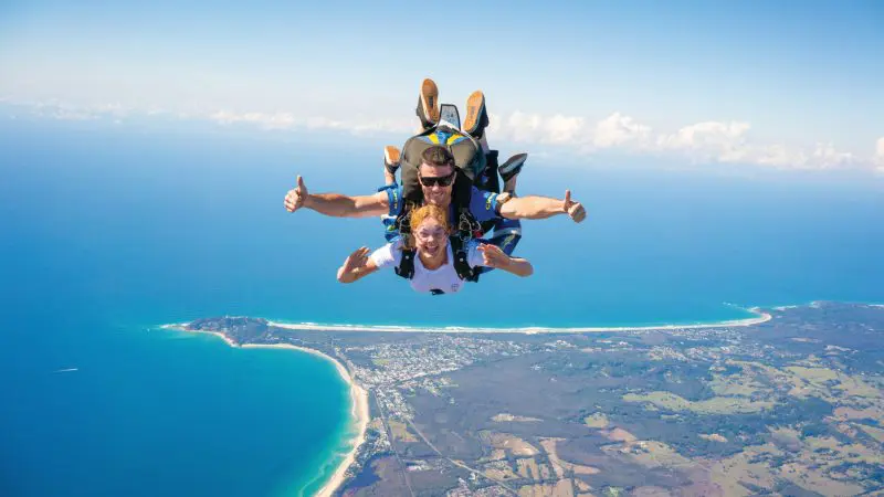 Adventurous tandem skydivers at Skydive Byron Bay smiling and giving thumbs up high above Australia’s breathtaking coastal scenery.