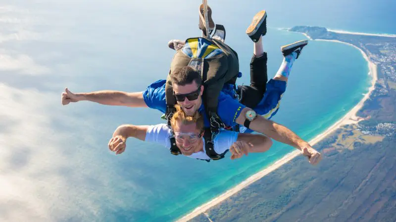 Tandem skydivers freefalling from 15,000ft over Byron Bay coastline, smiling above turquoise sea under clear blue sky.