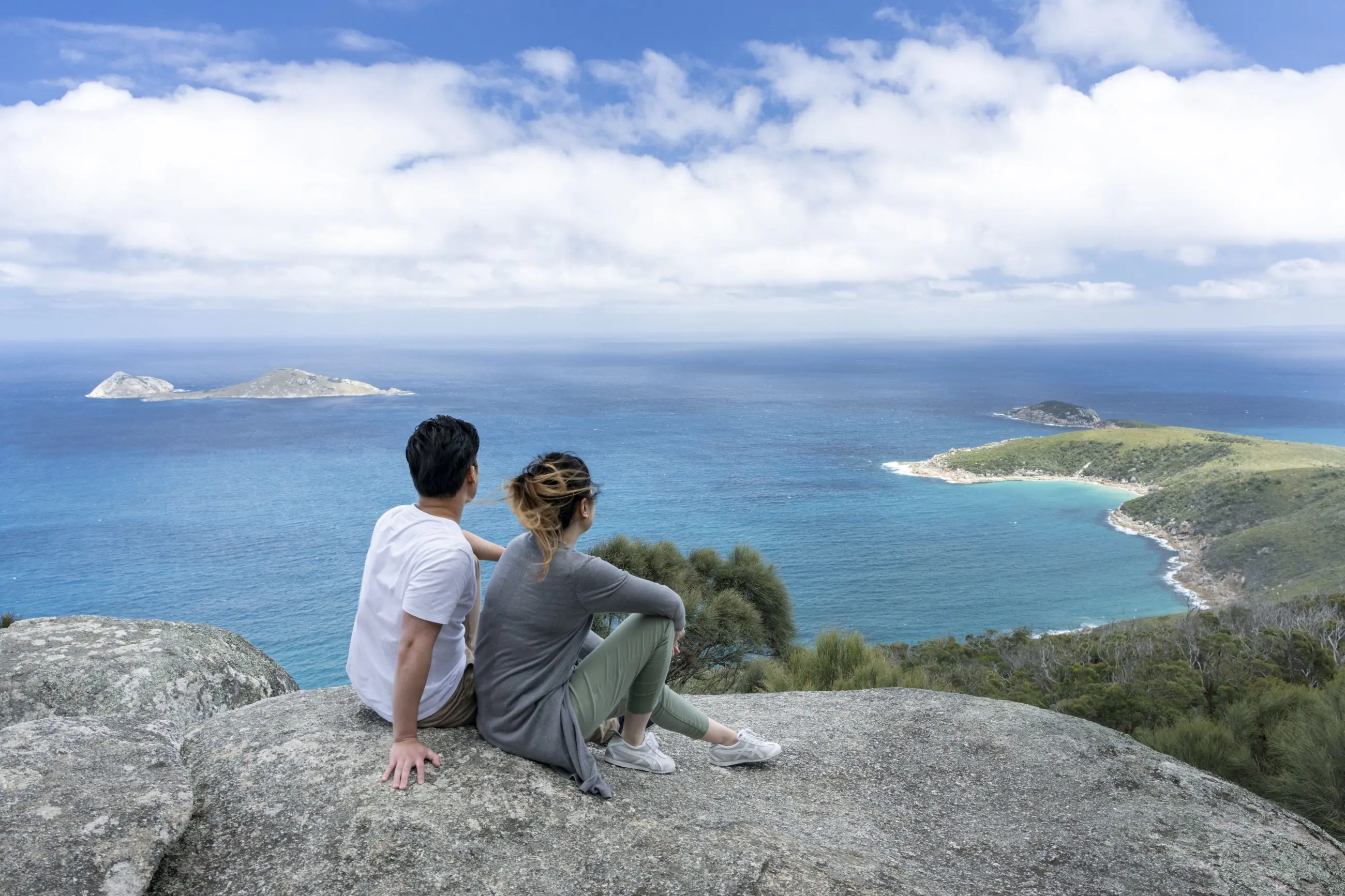 A couple relaxes atop a rocky cliff at Wilsons Promontory Day Tour, overlooking scenic sea views, islands, and lush coastline.