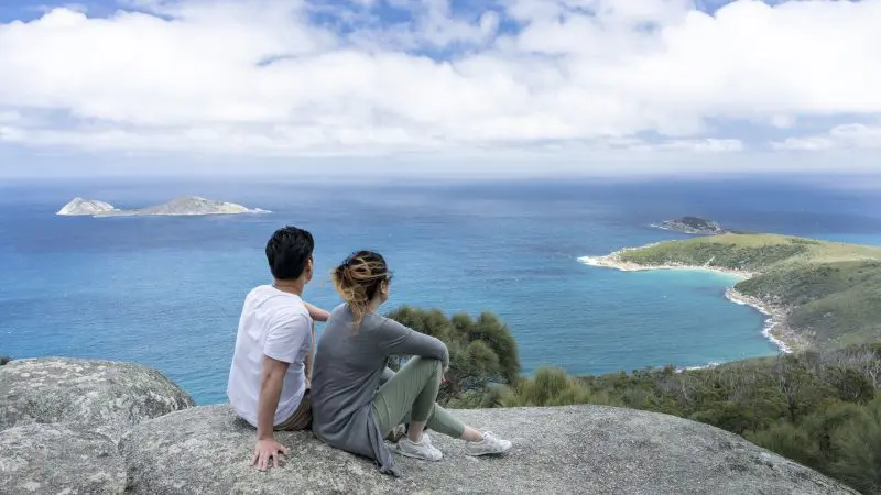 A couple relaxes atop a rocky cliff at Wilsons Promontory Day Tour, overlooking scenic sea views, islands, and lush coastline.