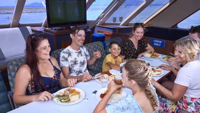 Happy family enjoys a meal and laughter aboard a Full Day Green Island 11AM departure boat, with social distancing sign visible.
