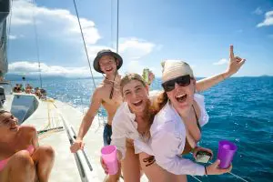Three friends smiling on a sailboat during their 28 Day Oz East Coast Adventure, enjoying drinks and stunning ocean views in the background.