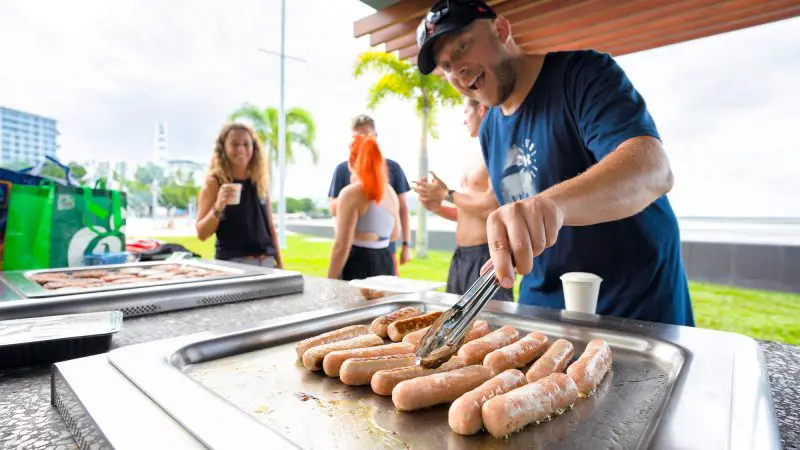 Man barbecuing sausages outdoors at 28 Day Oz East Coast Adventure Intro Travel event, highlighting group travel experience.