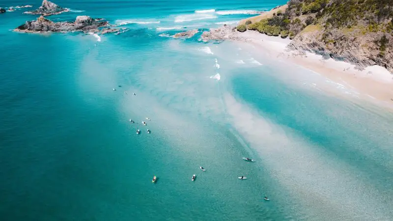 Stunning aerial shot of turquoise sea, kayakers, golden sandy beach, and rugged rocky shore on 10 Day Oz Intro Sydney to Brisbane tour.