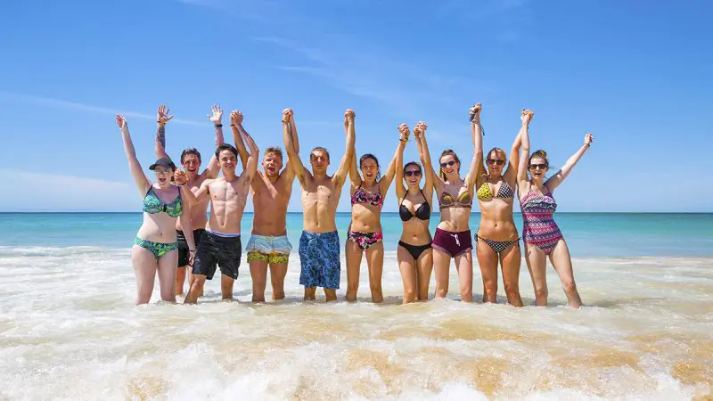 Group enjoying 10 Day Oz Intro Sydney to Brisbane tour, standing in sea waves, smiling and posing under clear blue sky for photos.