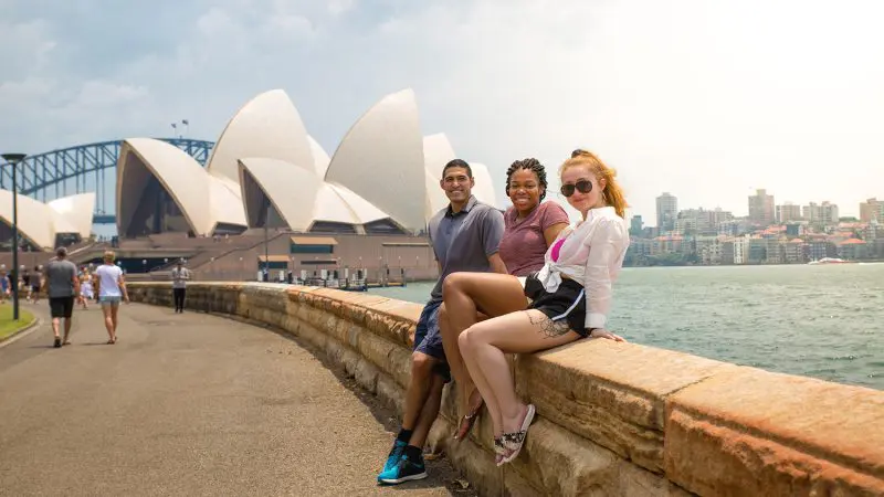 Three friends enjoy waterfront views on a stone wall during their 10 Day Oz Intro Sydney to Brisbane adventure travel experience.