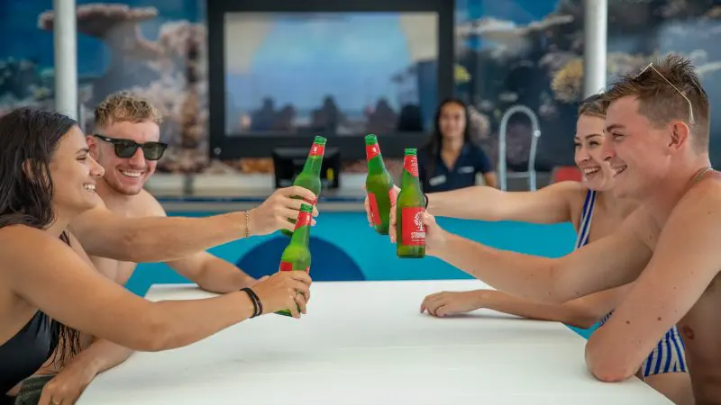 Group of friends in swimwear toasting with green beers on Reef Magic Outer Barrier Reef Pontoon, enjoying a vibrant tropical holiday.
