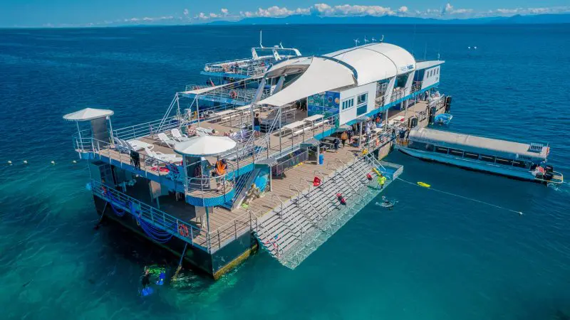 Aerial view of Reef Magic Outer Barrier Reef Pontoon with swimmers, boats, and tourists in crystal-clear blue water under sunny skies.