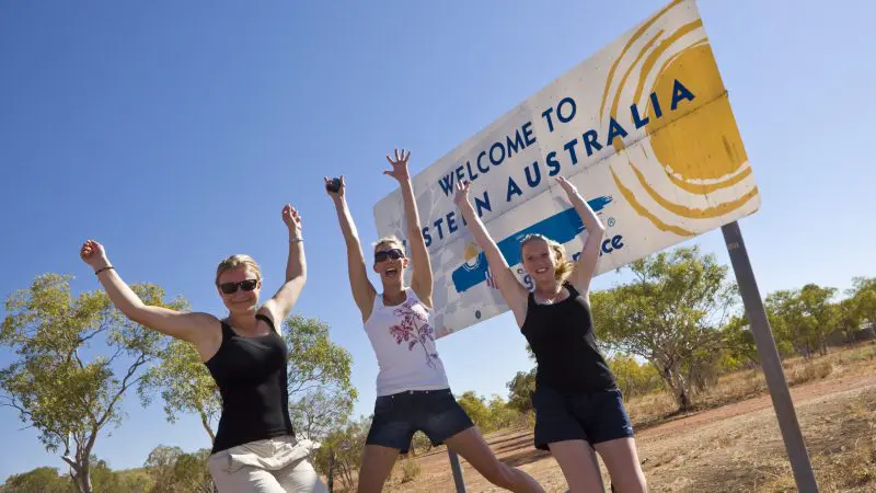 Three happy travellers jump and smile by a 'Welcome to Western Australia' sign—Perth to Darwin tour package includes Broome stays.