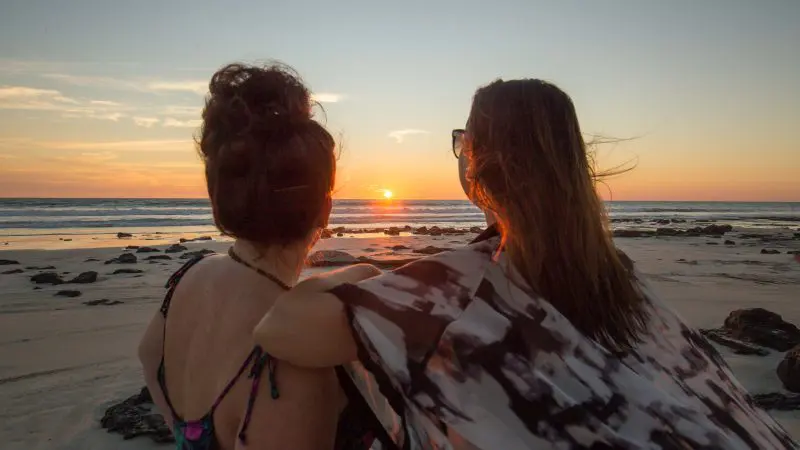 Two women watch a vibrant sunset on a rugged beach, envisioning their unforgettable 10 Day Broome to Darwin 4WD Kimberley Adventure.