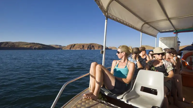 Tourists enjoy a scenic boat ride on a pristine lake surrounded by rocky hills under clear skies during a 10 Day Broome to Darwin Kimberley 4WD Adventure.