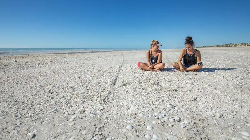 Two women enjoy a shell-strewn beach, smiling and relaxing on their Darwin to Perth Adventure Combo trip under sunny skies.