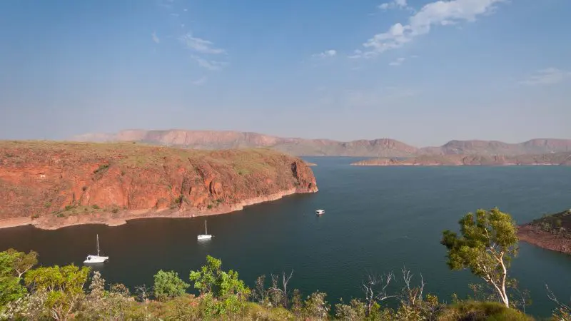 Expansive river flanked by striking red cliffs and rolling hills, dotted with sailing boats—evoking a top-rated Darwin to Perth tour.