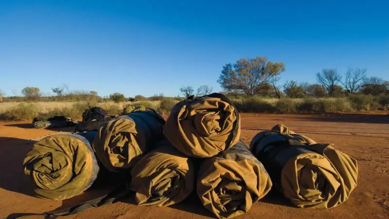 Six rolled-up sleeping bags on rich red outback soil, prepared for a two-night MacDonnell Ranges adventure from Alice Springs.