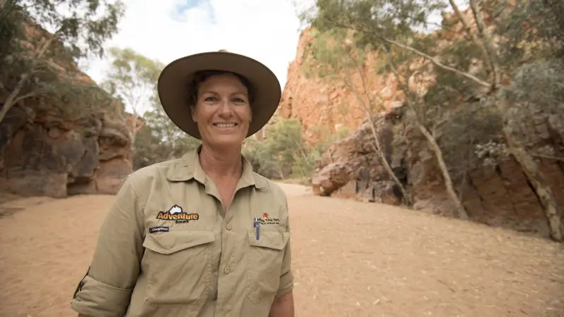 Smiling woman in hiking gear treks through rugged outback canyon on 5-day camping tour from Yulara to Alice Springs, Australia.