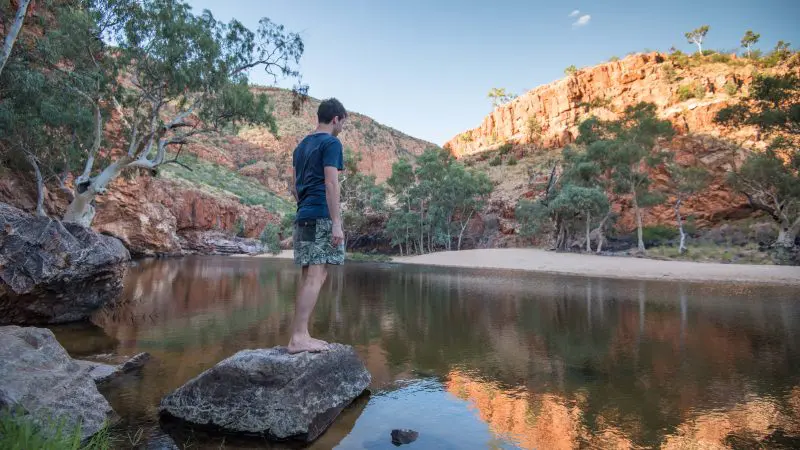 Barefoot man on a rock beside a tranquil river in the MacDonnell Ranges at sunset, enjoying his 2-night adventure tour.