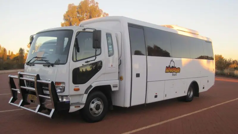 White Adventure Bus for 2 Day 1 Night Uluru Adventure parked on red brick pavement in Yulara, surrounded by lush green trees.