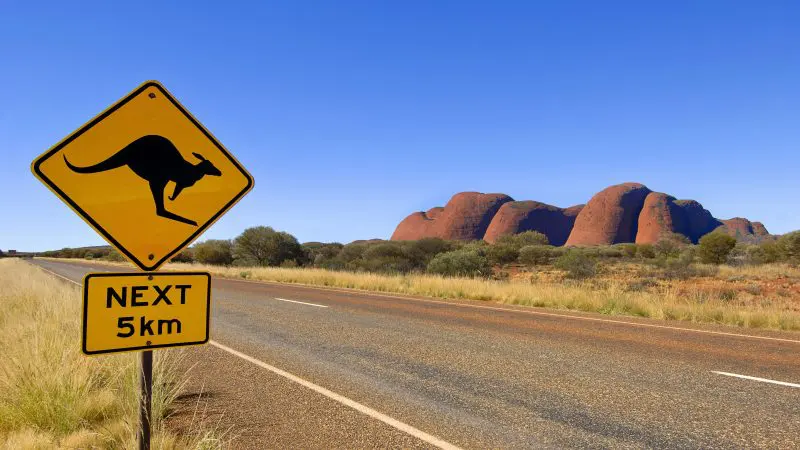 Bright yellow kangaroo road sign with "NEXT 5km" on scenic Uluru tour, Yulara loop; iconic red rock formations in the background.
