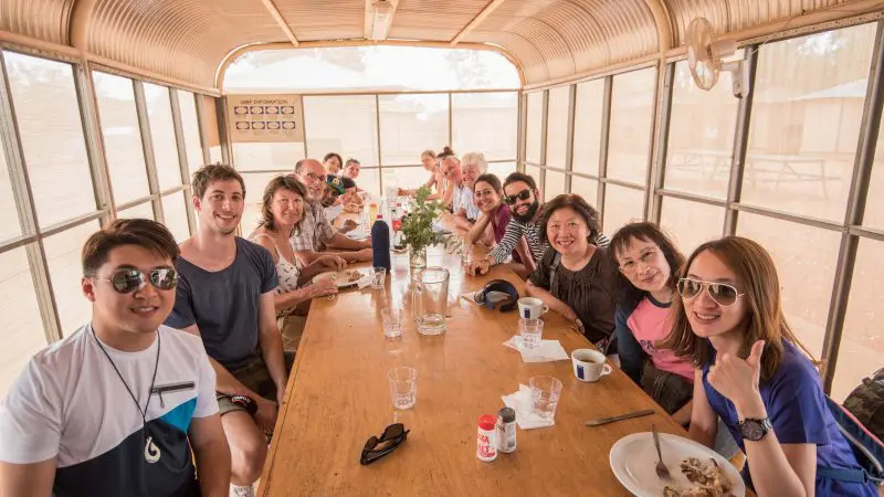 Happy travellers gather around a rustic wooden table, sharing stories during a 2 Day 1 Night Uluru Adventure from Yulara.