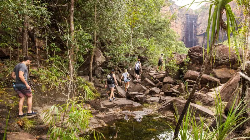 Adventurers trek across rocky terrain in dense, green forest beside a waterfall during a 4 Day Kakadu, Katherine & Litchfield tour.
