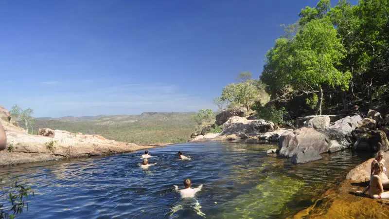 Adventurers swim in a scenic Kakadu rock pool, encircled by lush trees and rugged rocks on a 4WD tour beneath vibrant blue skies.